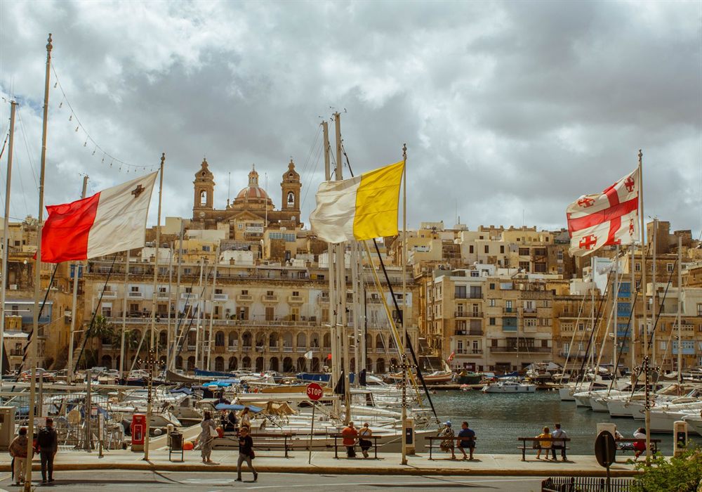 Harbor in Malta lined with boats under flags, with historic stone buildings behind